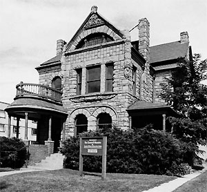 A black and white photo of the mansion with a sign in the foreground and foliage on the base and side. 