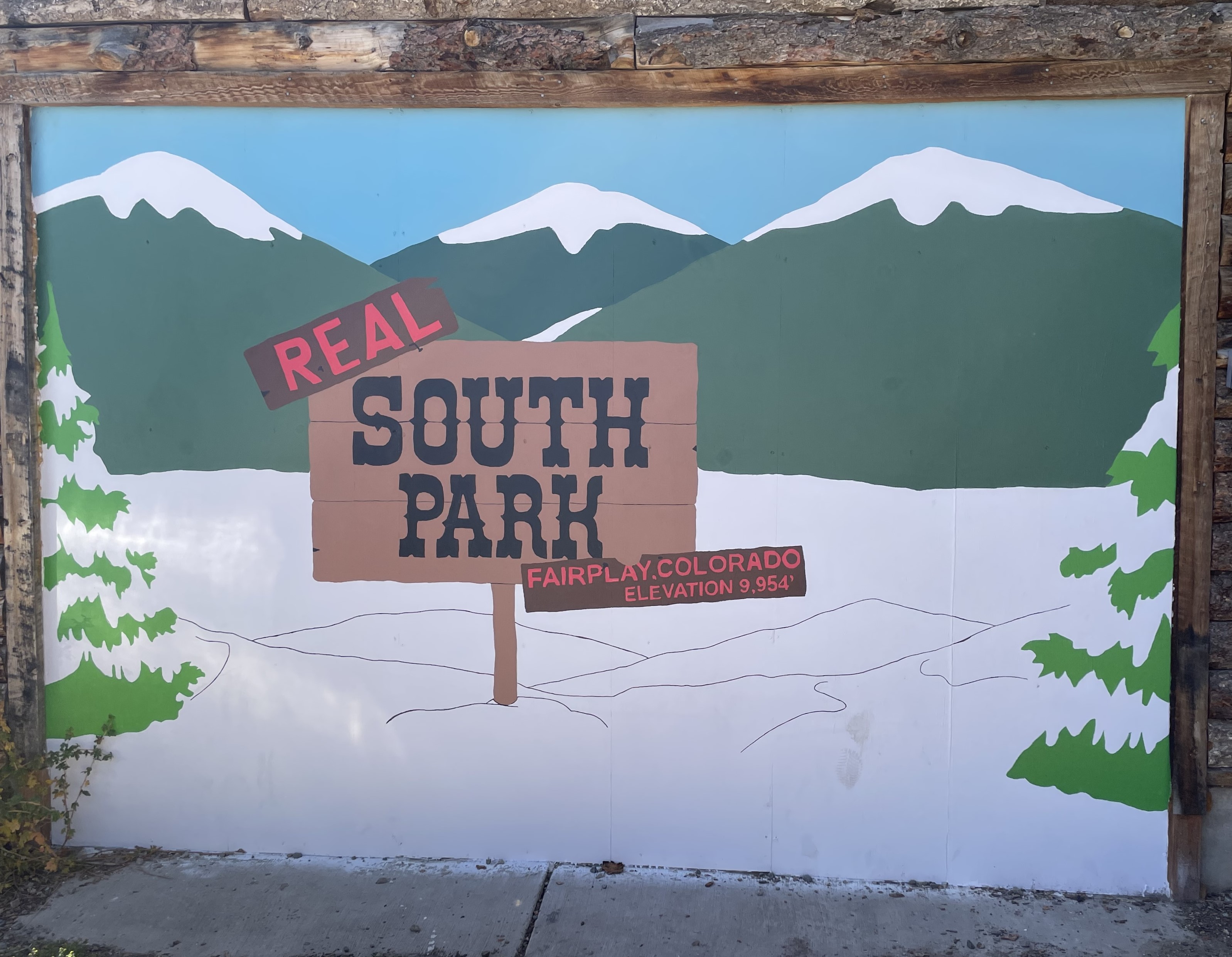 2024 Photograph of South Park Mural in Fairplay. In the style of South Park animation, the mural shows a wooden sign in the snow that reads, Real South Park: Fairplay, Colorado, Elevation 9,954’.  There are snow covered fir trees on either side of the sign and green mountains with snowy tops in the background. 