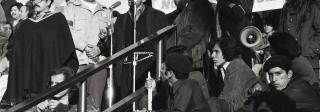 Photo of an outdoor anti-war protest rally featuring speaker Corky Gonzales. He stands in front of a microphone flanked by unidentified men and women standing and sitting on the steps. A banner behind Corky reads "RAZA SI, GUERRA NO."