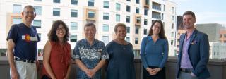 A group of six individuals stand on a balcony in Denver, with the city skyline and cranes behind them.