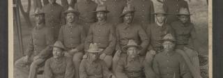 Black and white photo of soldiers in uniform standing or sitting in rows in front of trees.