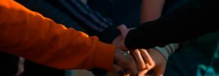 Photograph of a group of people who are stacking up their hands as if in a team going to play a game, or a group of people getting ready to undertake a team task. The sun shines on their hands, and several of the people wear long-sleeved colorful clothing.
