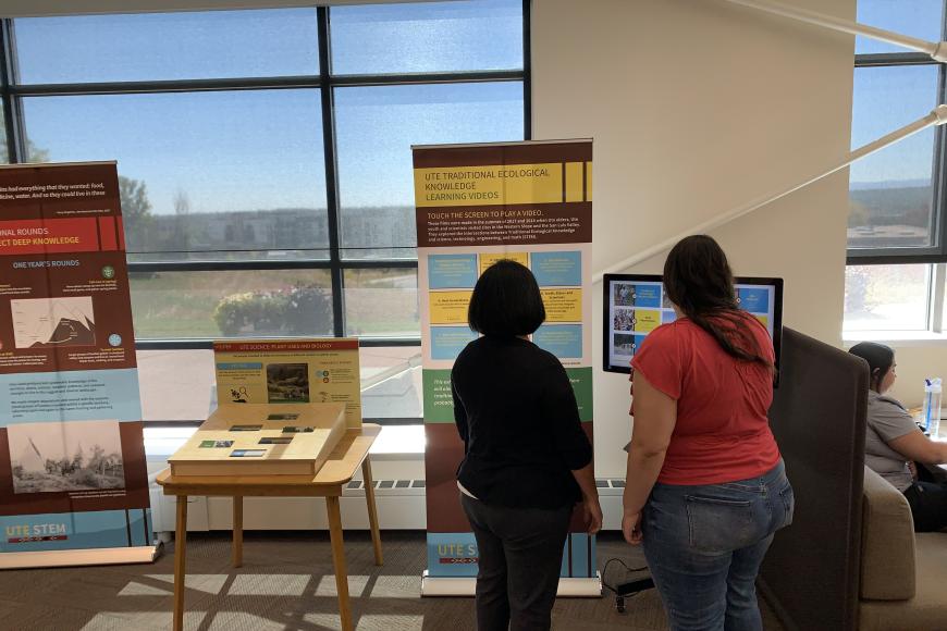 Two women reading a pop-up panel about Ute knowledge, in front of a large window.