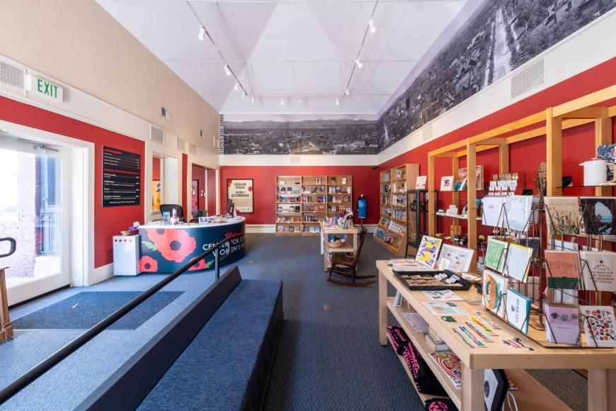 Interior of Center for Colorado Women's History gift shop. It has red walls, and open interior with tables lined with books, accessories, and other gifts.