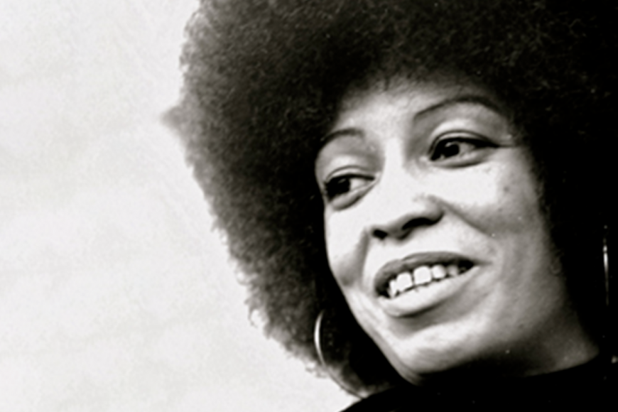 Angela Davis in a black turtleneck, overcoat, and grey sweater with hoop earrings is seen smiling away from the camera. 
