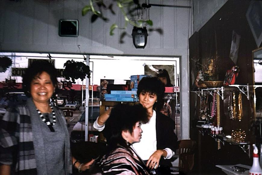 3 women, 2 hairdressers and a customer, smile while styling a dramatic 80s haircut.