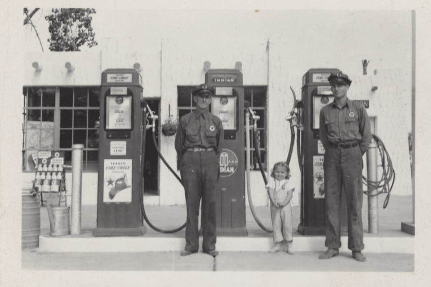 Two police officers and a young girl stand in front of vintage gas pumps.