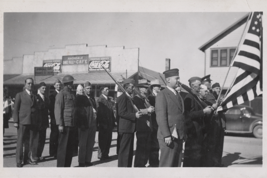 	 Black and white photograph of World War One veterans marching down the street. The men are carrying the US flag, and some are wearing their military uniform. There are shops in the background.