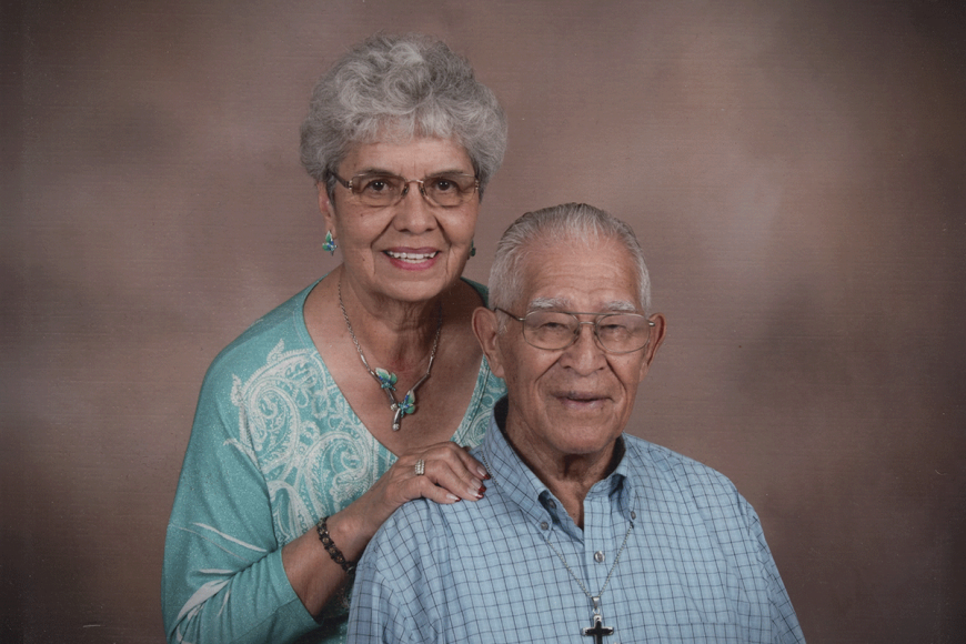 An older couple poses for a studio photograph. The woman is leaning over the man's shoulder and smiling.