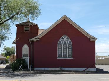 The photo is of a brick church painted red. To the left side of the image there is a two story tower that marks the entrance to the building. To the right of the tower is the south elevation of the church. It contains a pointed arch window in the center of the elevation with stained glass inserts. 