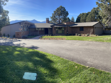 Pictured is the facade of the Hill Summer Residence. In front of the residence is a driveway and lawn. The house is a single-story, mid-century ranch house. 