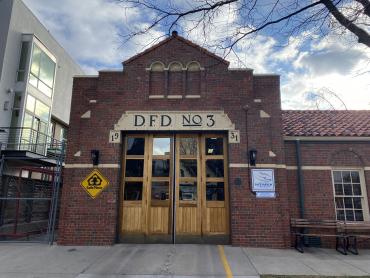 In the picture is the northern portion of the west elevation of the Fire Station. This portion has garage doors for the single-engine room including two wooden bi-fold garage doors. Each section of the door has one row of three vertical glass panes on the top section, a vertical boarding lower section, and a metal kick plate. There is decorative white stonework above and around the top edge of the doors. 