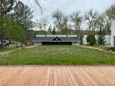 Photo is of the Boulder Kyudojo. There is wood platform at the forefront of the photo with a long green space in front of that. Behind the green space is the building that houses the archery range. 