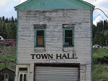 Photo is of the Red Cliff Town Hall. A two story building with a gable front roof. There is a large garage door on the first floor with a door to the left. The second floor has a pair of vertically oriented rectangular windows that are centered on the facade. 