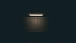 Photo of one single long flourescent light bulb, mounted to the ceiling of a dark area such as an empty parking garage. The light is illuminated, casting a glow over one concrete wall. The rest of the area remains dark.