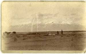 Sepia-toned historical photograph of men and horses in front of a snow-capped mountain range